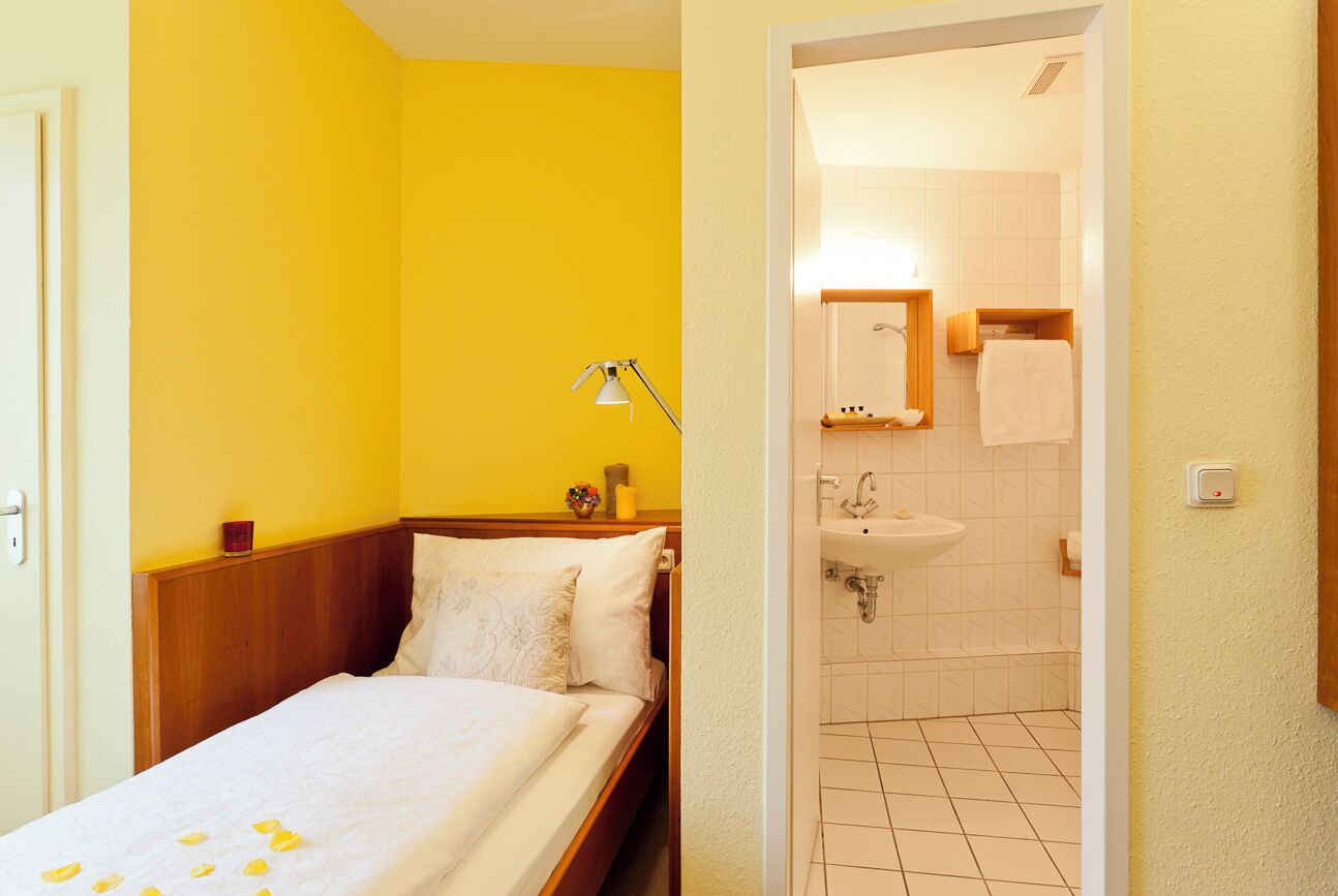 Une chambre d’hôtel avec un lit, une lampe de chevet, une petite table sur laquelle se trouve un vase, et une salle de bain visible à travers une porte ouverte.