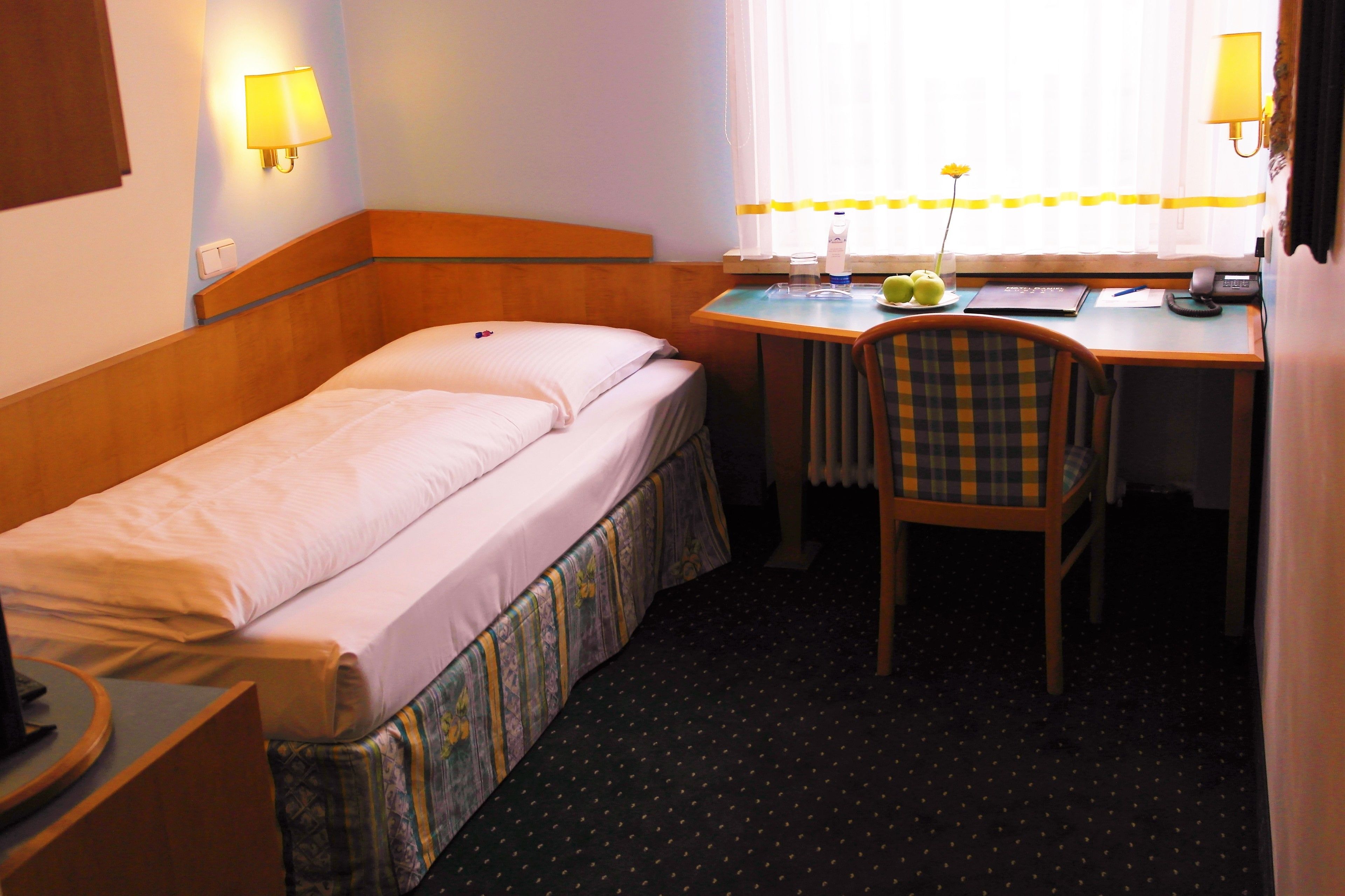 A small hotel room with a single bed, a desk, a chair, and a window with curtains.