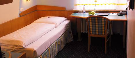 A small hotel room with a single bed, a desk, a chair, and a window with curtains.