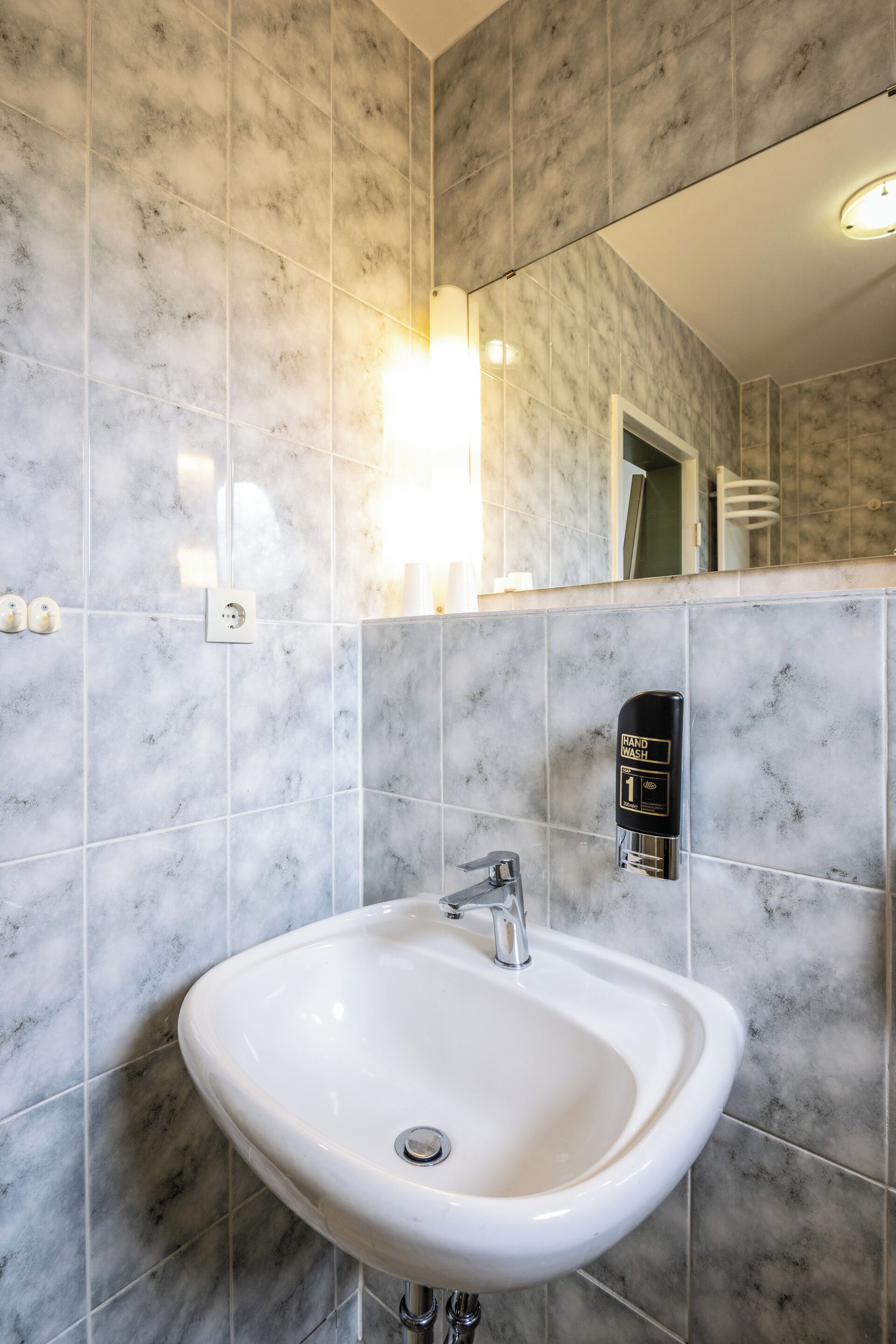 A bathroom with a white sink, marble tiles, a hand wash station, and a mirror.