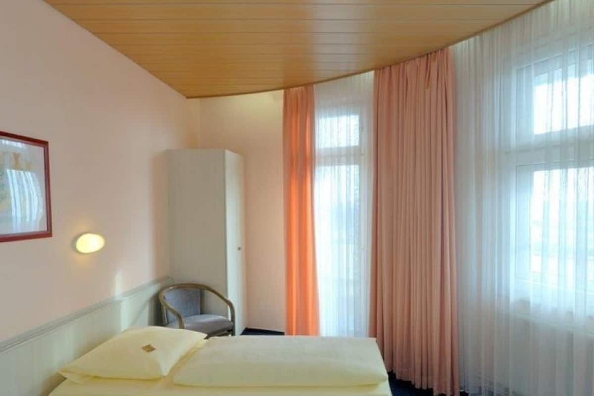 A hotel room with a bed, a chair, a wardrobe, and a window with curtains.