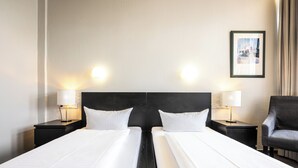 A hotel room with two single beds, a black headboard, bedside lamps, a framed picture on the wall, and a grey armchair. - Raststättenhotel Avus (Berlin)
