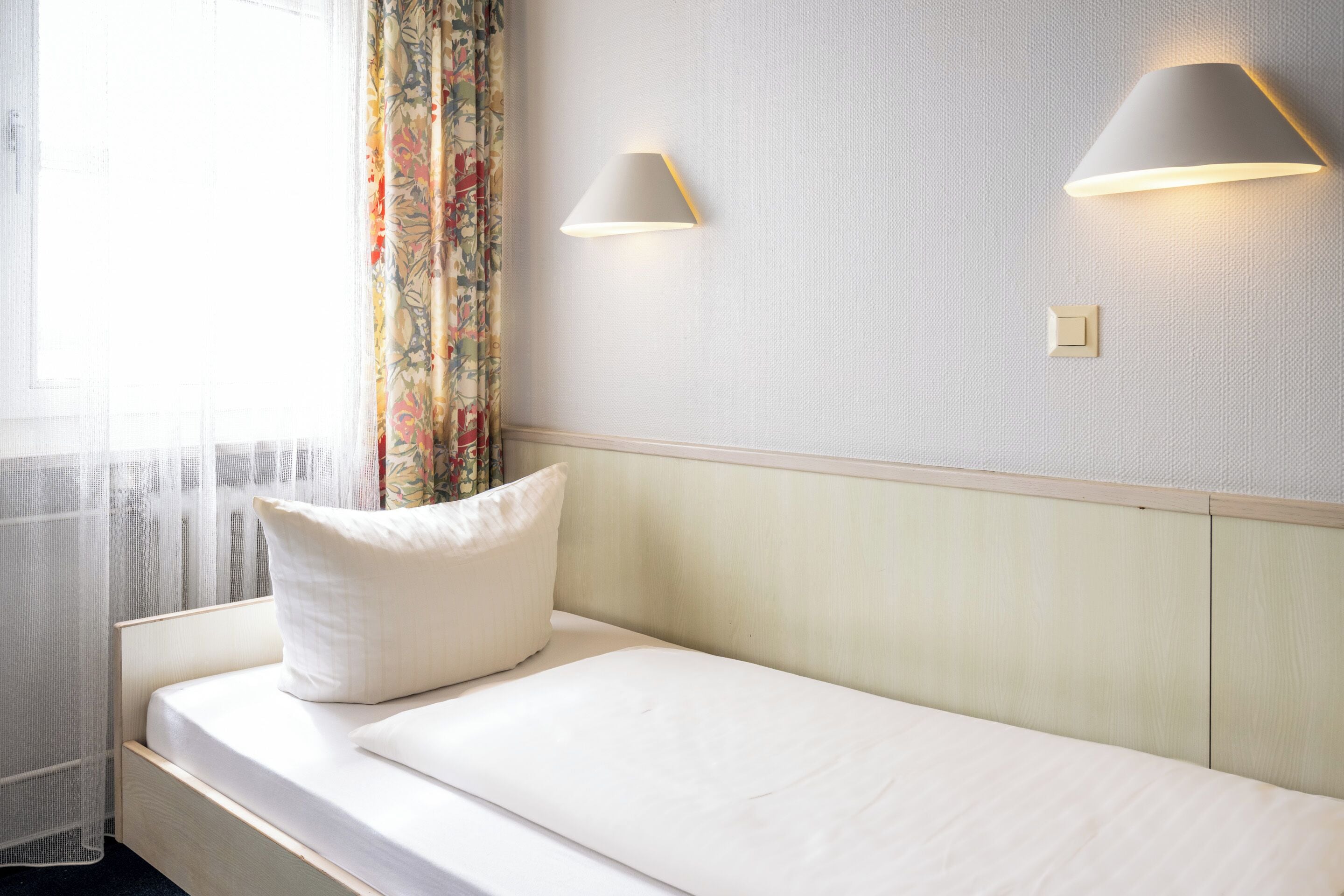 A single bed with a white mattress and pillow, a window with sheer curtains, and two wall-mounted lamps.