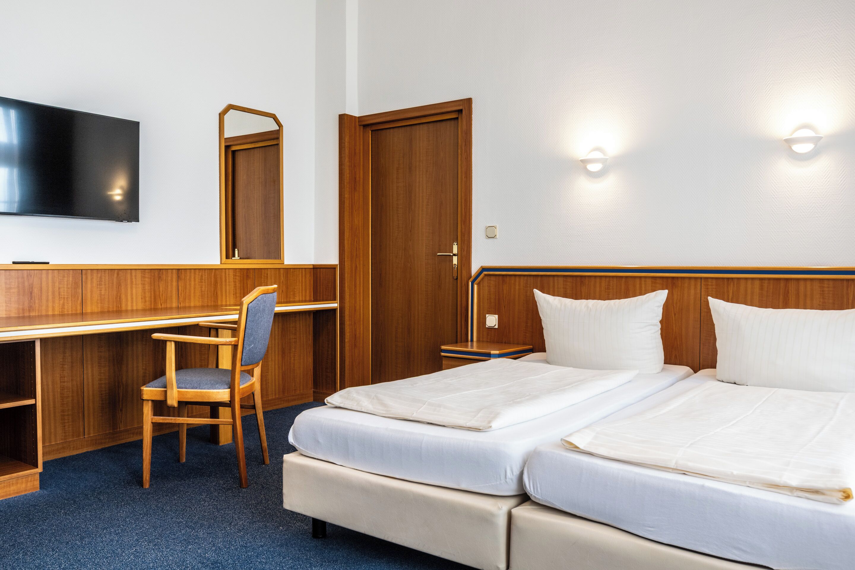A hotel room with a desk, a chair, a bed with two pillows, a TV, and a door.