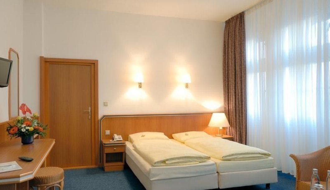 A hotel room with a double bed, bedside tables, a desk with a chair, a TV, and a window with curtains.