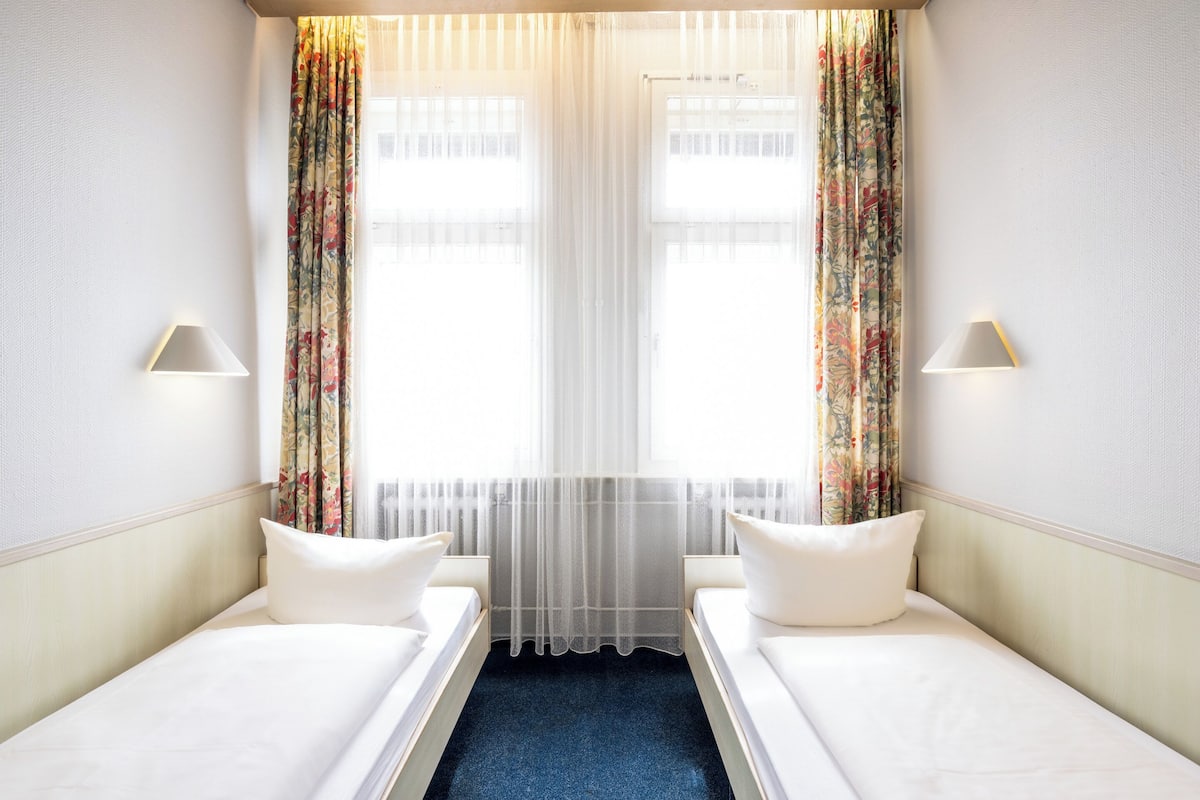 A hotel room with two single beds, a window with sheer curtains, and two wall-mounted lamps.