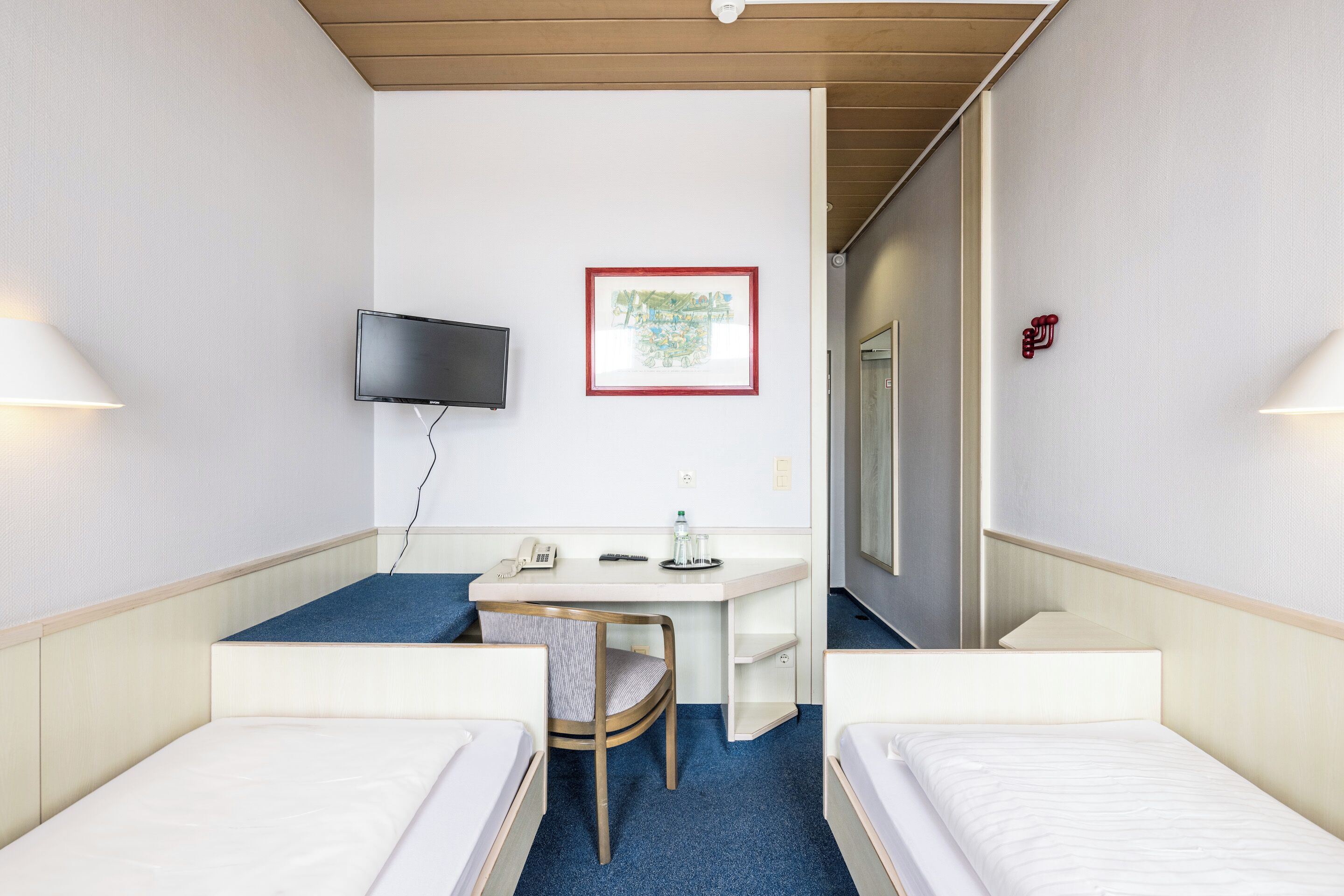 A hotel room with two single beds, a desk with a chair, a flat-screen TV mounted on the wall, a framed map, and a water bottle on the desk.