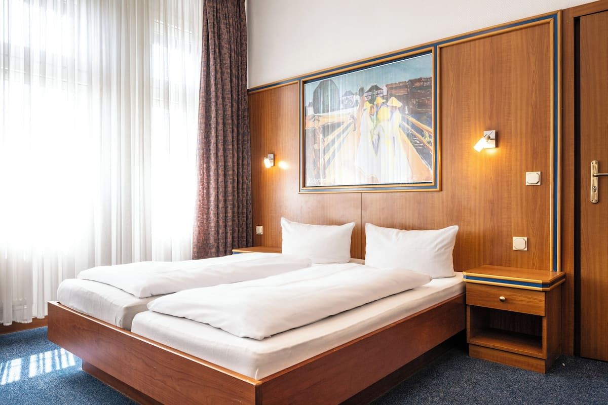 A double bed with white linens, a wooden headboard, a painting above the bed, and a nightstand with a lamp.