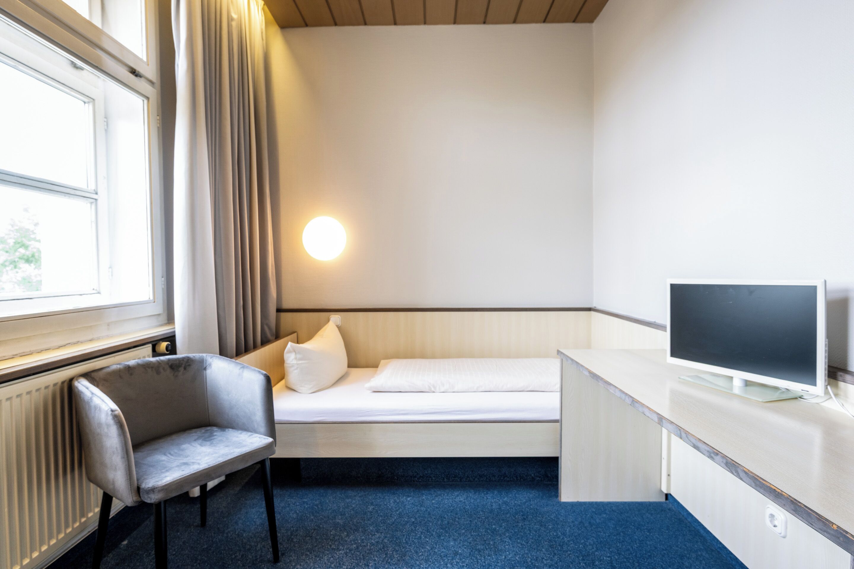 Una stanza piccola e ordinata con un letto, una scrivania, una sedia, una televisione e una finestra con tende.
