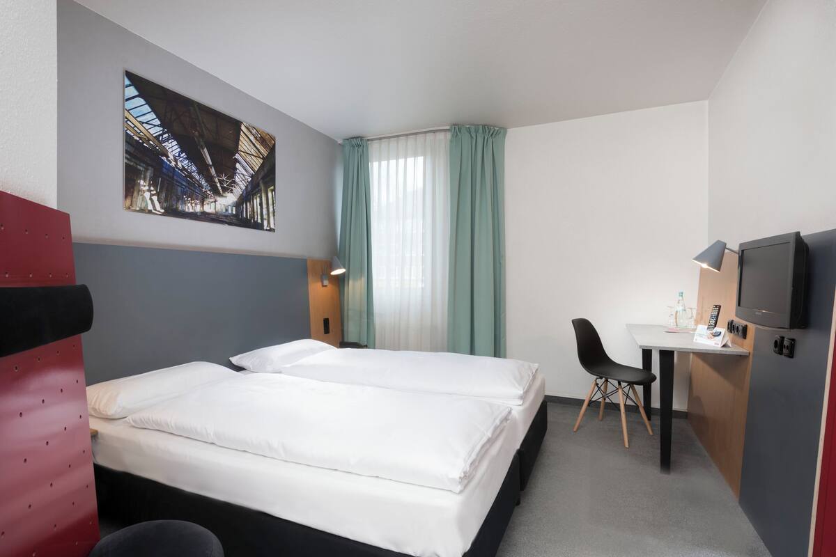 A hotel room with two beds, a desk, a chair, a TV, and a painting on the wall.