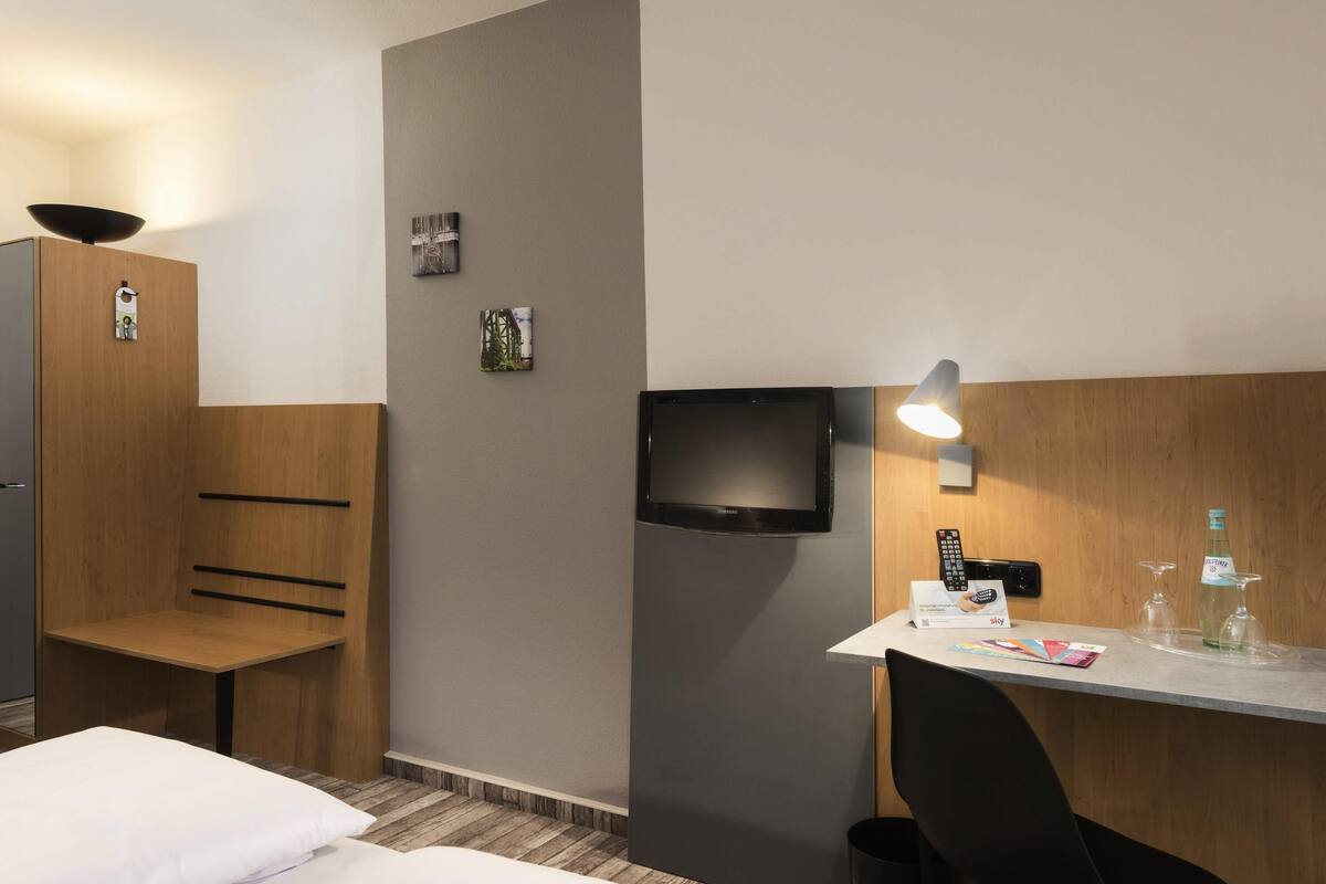A hotel room with a bed, a desk with a chair, a television, and a small table with a bottle and glasses.