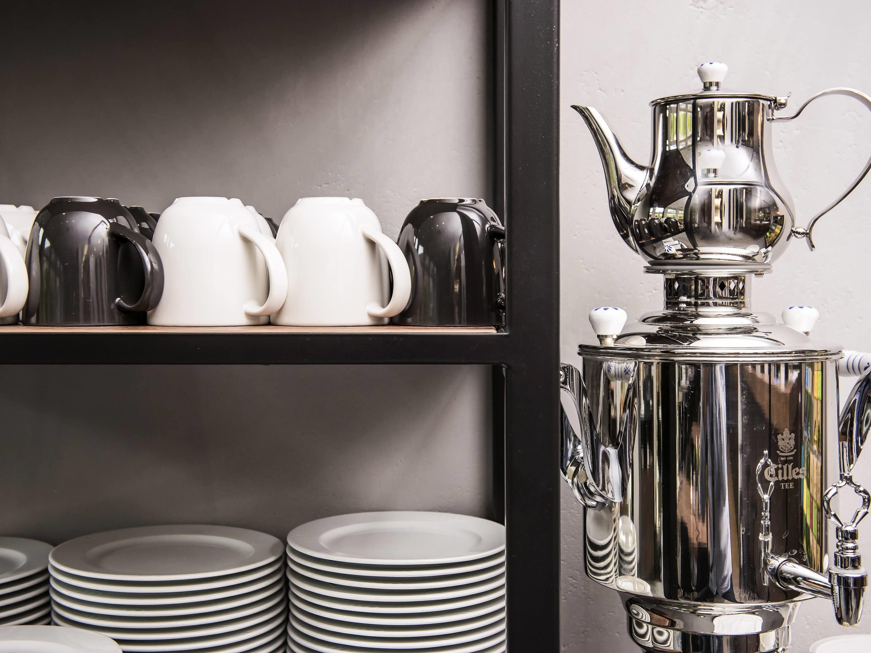 A kitchen area with a coffee station featuring a large silver teapot and multiple cups.