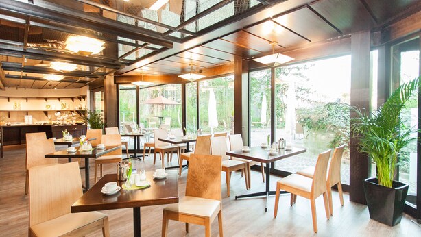 Un café spacieux avec des tables et des chaises en bois, de grandes fenêtres donnant sur un paysage verdoyant et un design moderne au niveau du plafond.