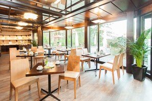 Un café spacieux avec des tables et des chaises en bois, de grandes fenêtres donnant sur un paysage verdoyant et un design moderne au niveau du plafond.