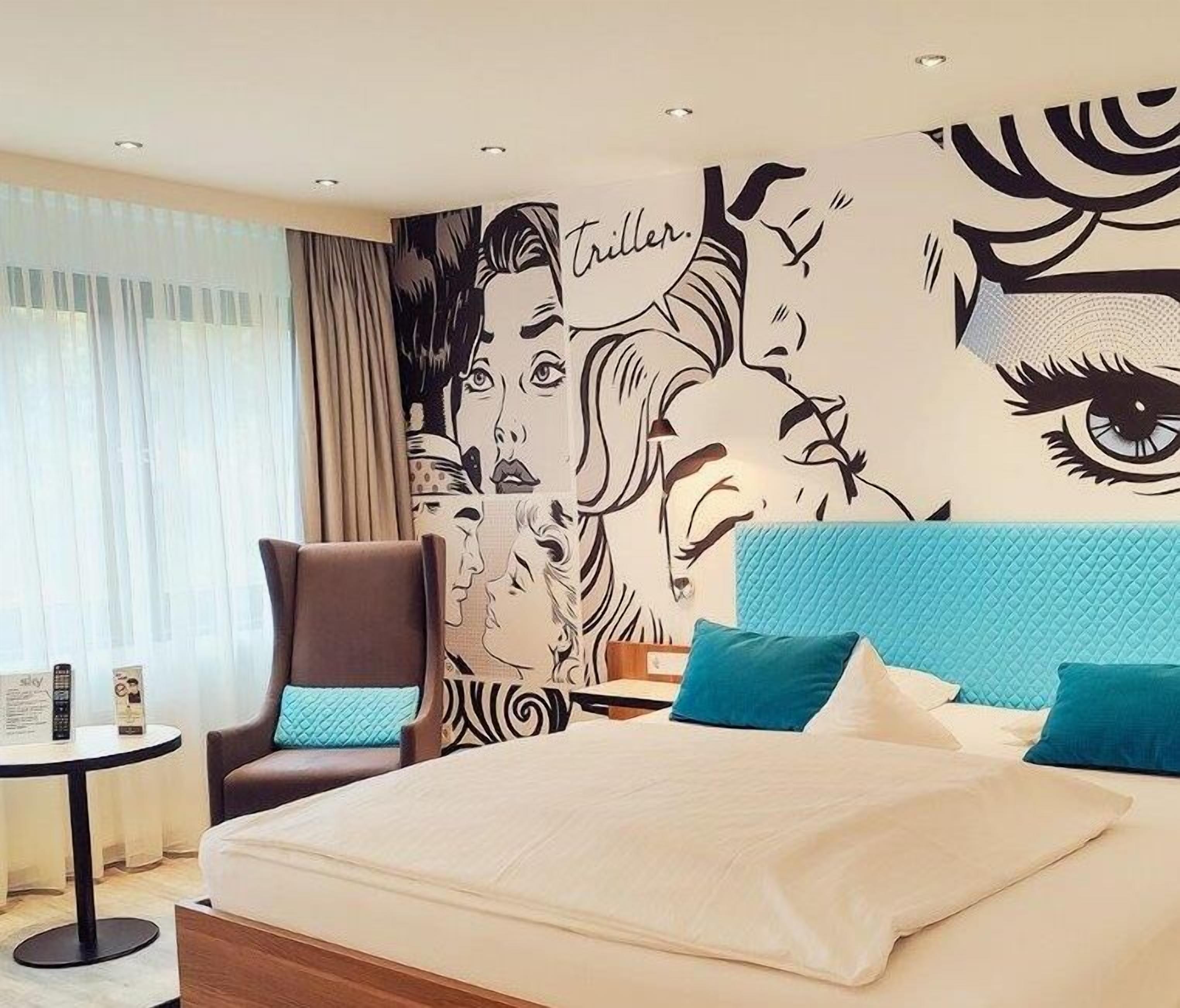 A hotel room with a large bed, a chair, a small table, and a wall with a graphic design.