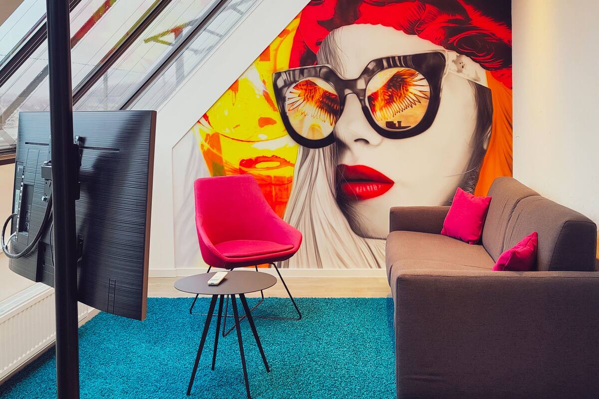 A modern living room with a vibrant mural, a brown sofa, a red chair, and a teal carpet.