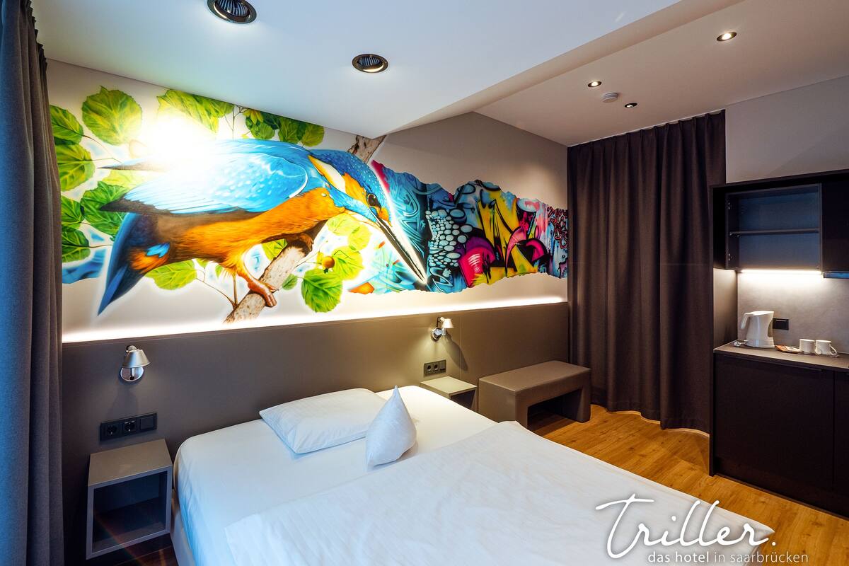 A modern hotel room with a large bed, a nightstand, a desk with a coffee maker, and a colorful mural on the wall.