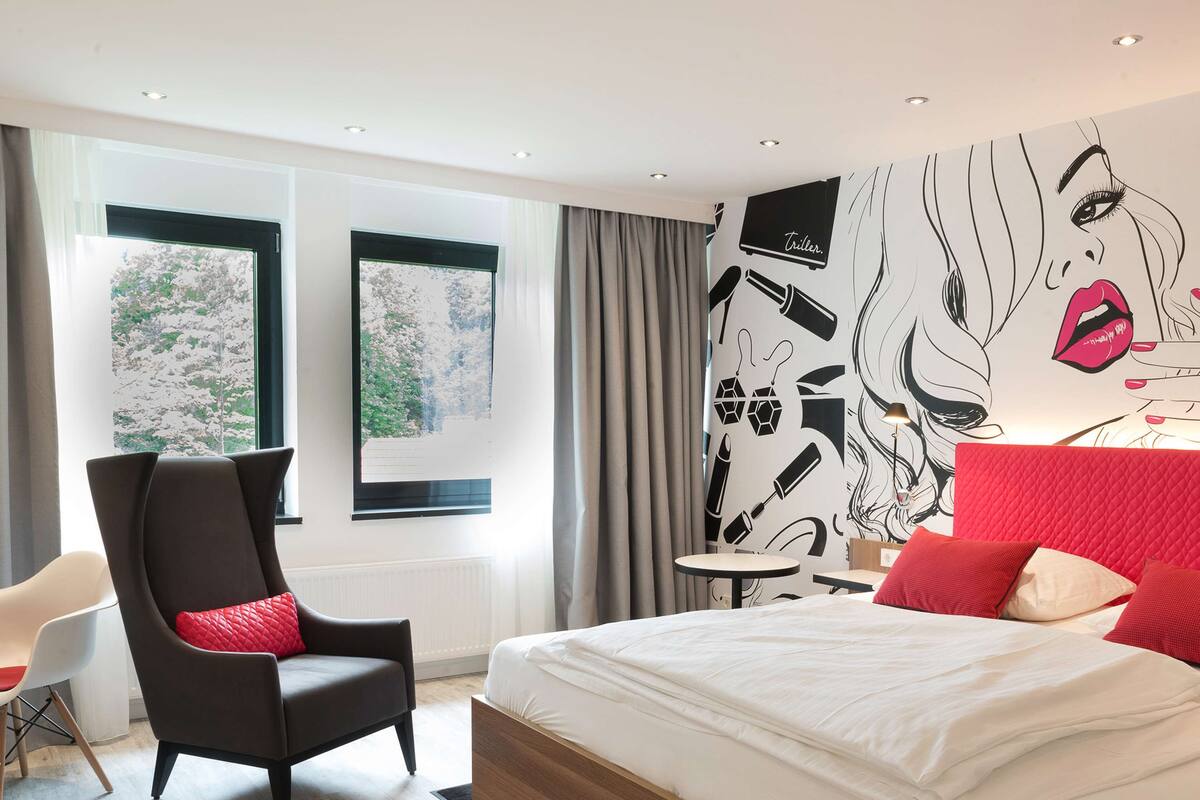 A modern hotel room with a large bed, a black armchair with a red pillow, a small round table, and a wall with a black and white abstract design.