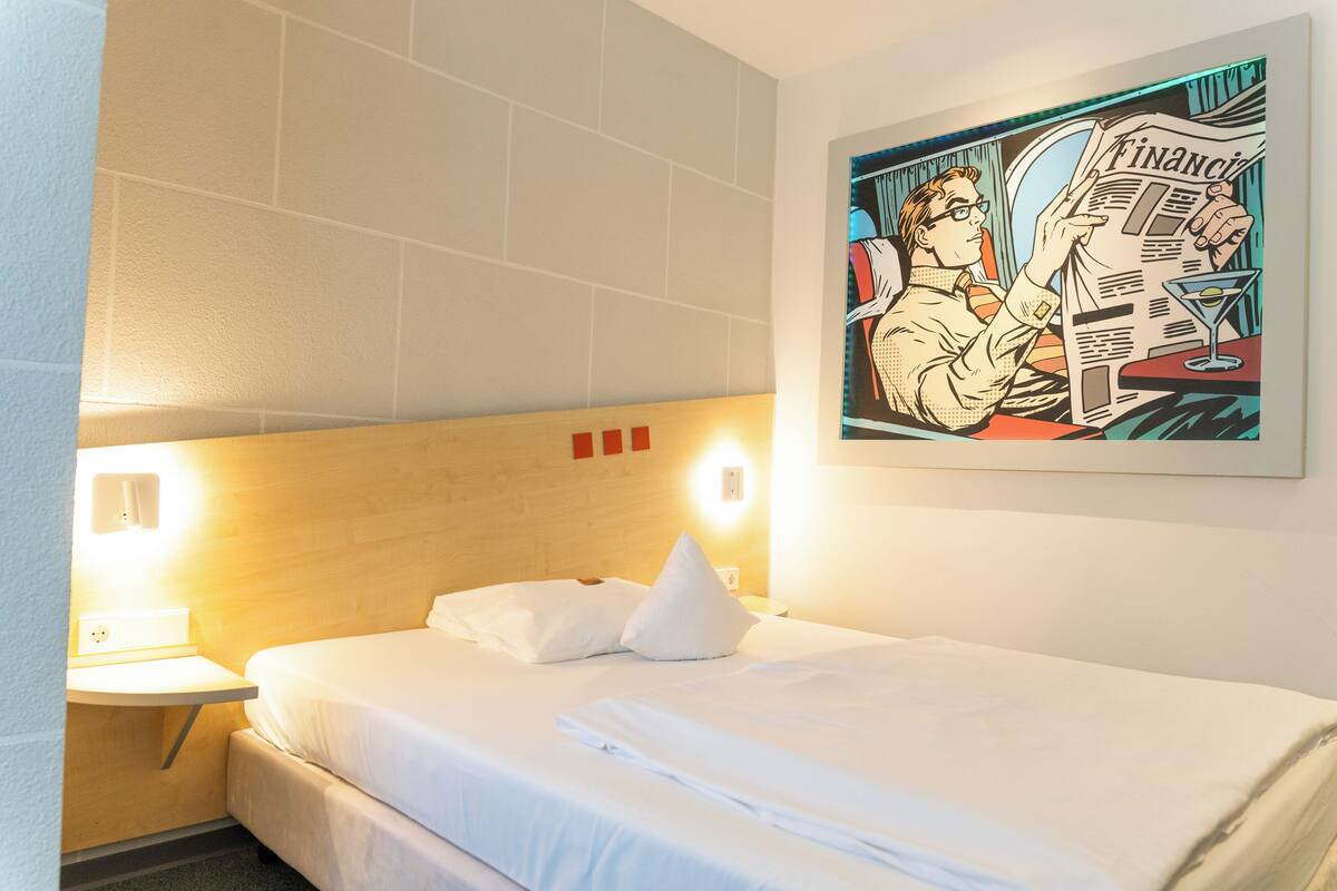 A hotel room with a bed, a nightstand, a wall-mounted light, and a poster of a man reading a newspaper.