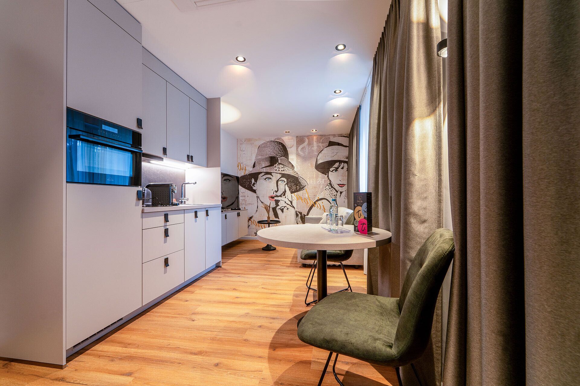 A modern kitchen with white cabinets, a built-in oven, and a round dining table with a green chair.