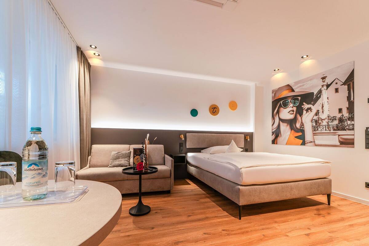 A modern hotel room with a bed, a sofa, a small table, and a wall-mounted TV.