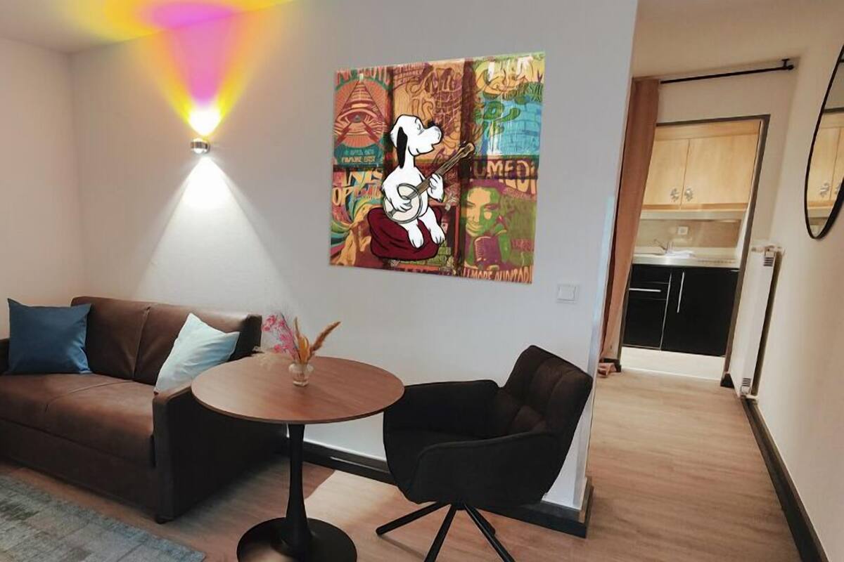 A living room with a brown sofa, a round table, and two black chairs. There are two wall art pieces, one with a pink character and another with a white character. A ceiling light is visible.