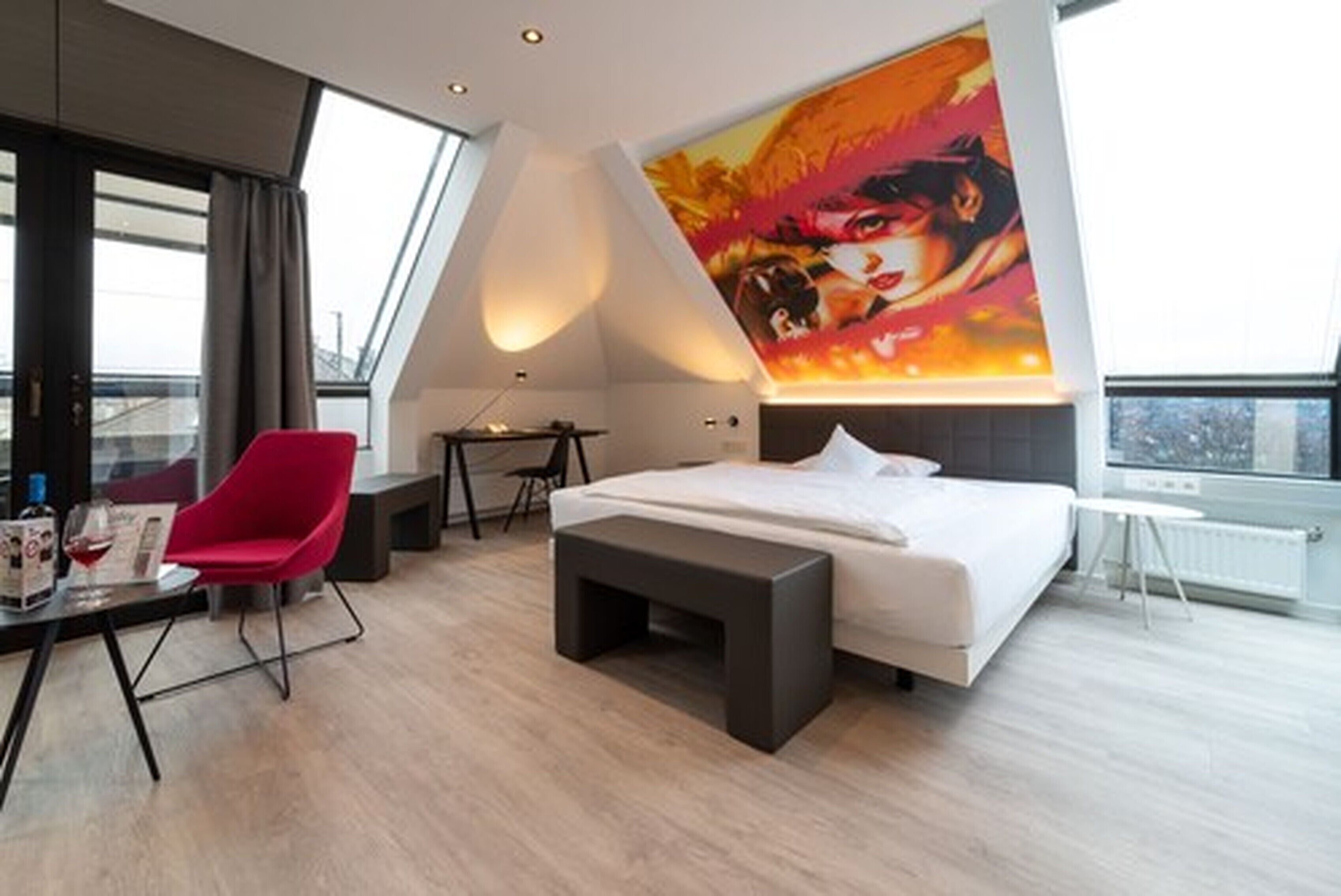 Een moderne hotelkamer met een groot bed, een rode stoel, een klein tafeltje met een fles en glazen, een bankje en een groot schilderij aan de muur.