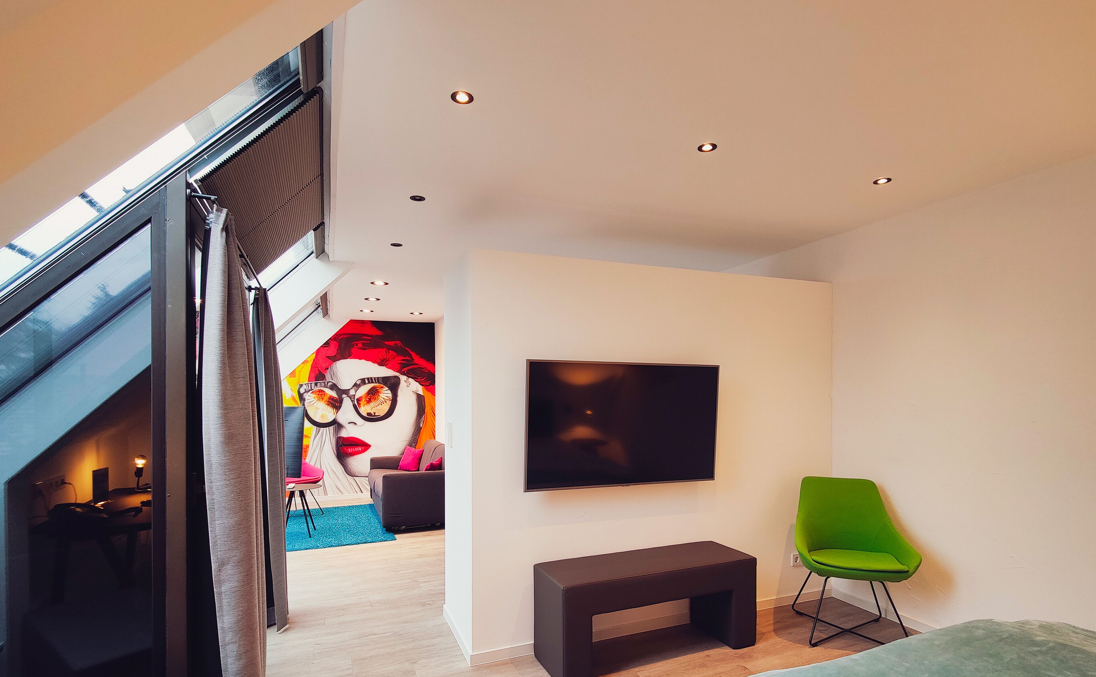 Una sala de estar moderna con un televisor de pantalla plana grande montado en la pared, un sillón verde, un banco y un mural colorido al fondo.