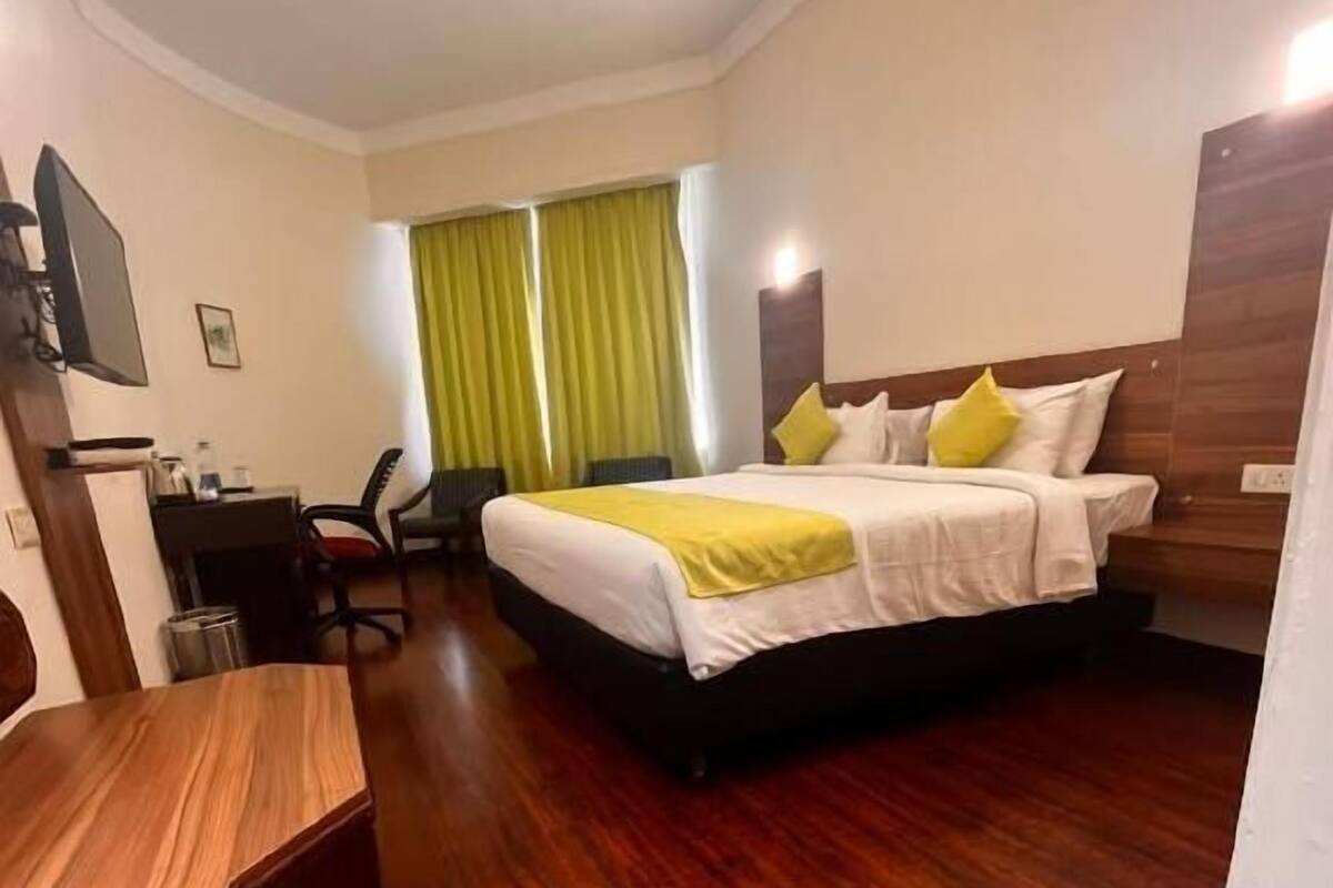 Habitación de hotel con piso de madera, cama con ropa blanca y almohadas amarillas, escritorio con silla y televisión en la pared.