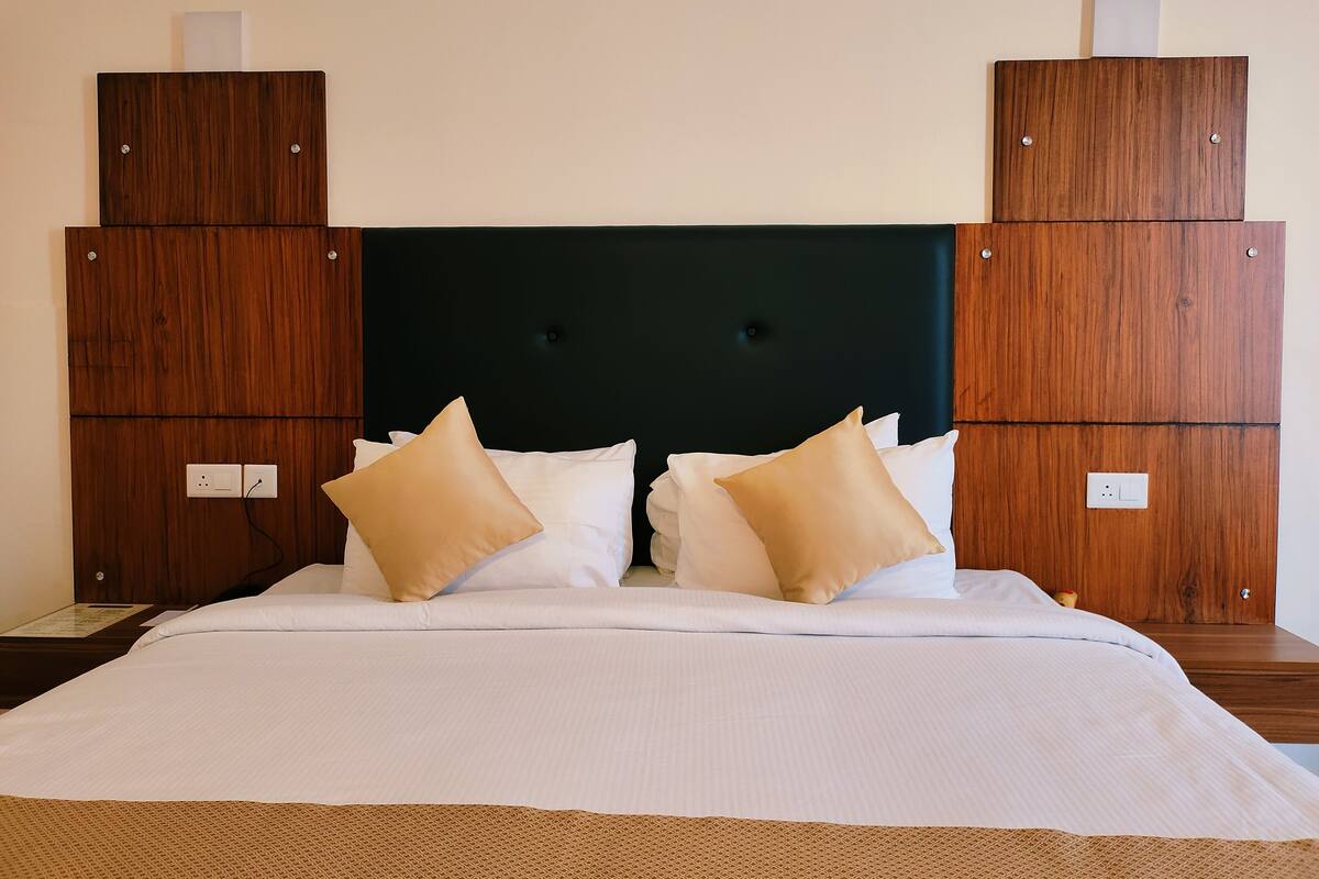 Una cama bien hecha con sábanas blancas y almohadas color beige, con dos mesitas de noche de madera, una de ellas con una pequeña planta.