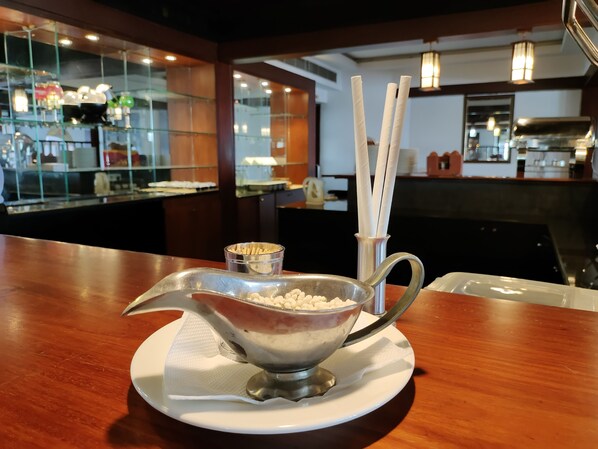 A dessert dish with a metal scoop on a white plate, a small bowl of nuts, and a container of white sticks on a wooden table in a restaurant.