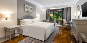 A hotel room with a large bed, a desk, a chair, a TV, and a view of greenery.