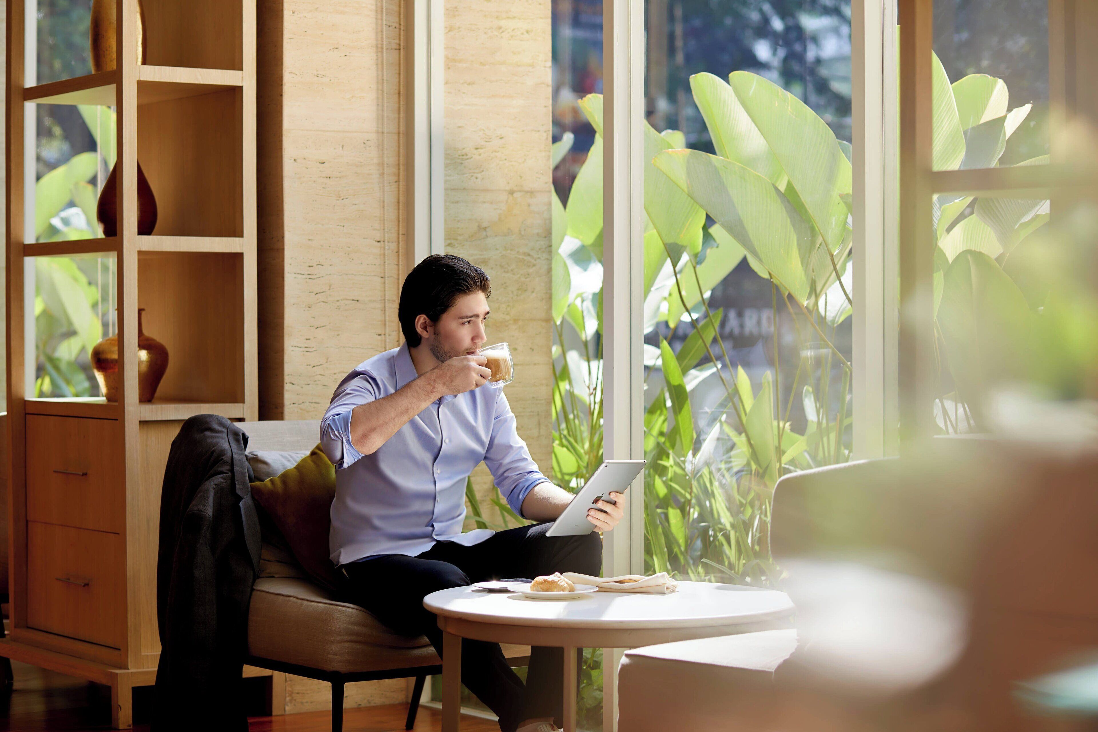 A man sitting at a table with a laptop and a glass of drink, in a room with a large window and a wooden cabinet.