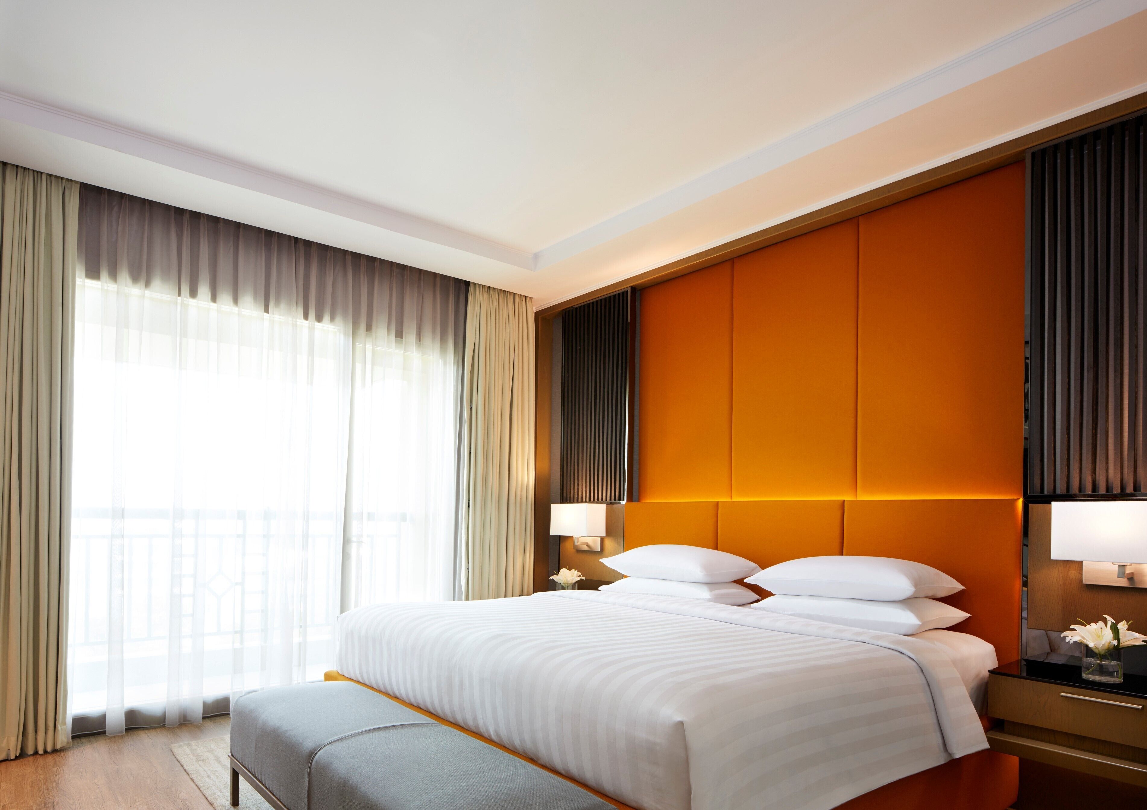 Een hotelkamer met een groot bed, een nachtkastje met een lamp, een raam met dunne gordijnen en uitzicht op buiten.