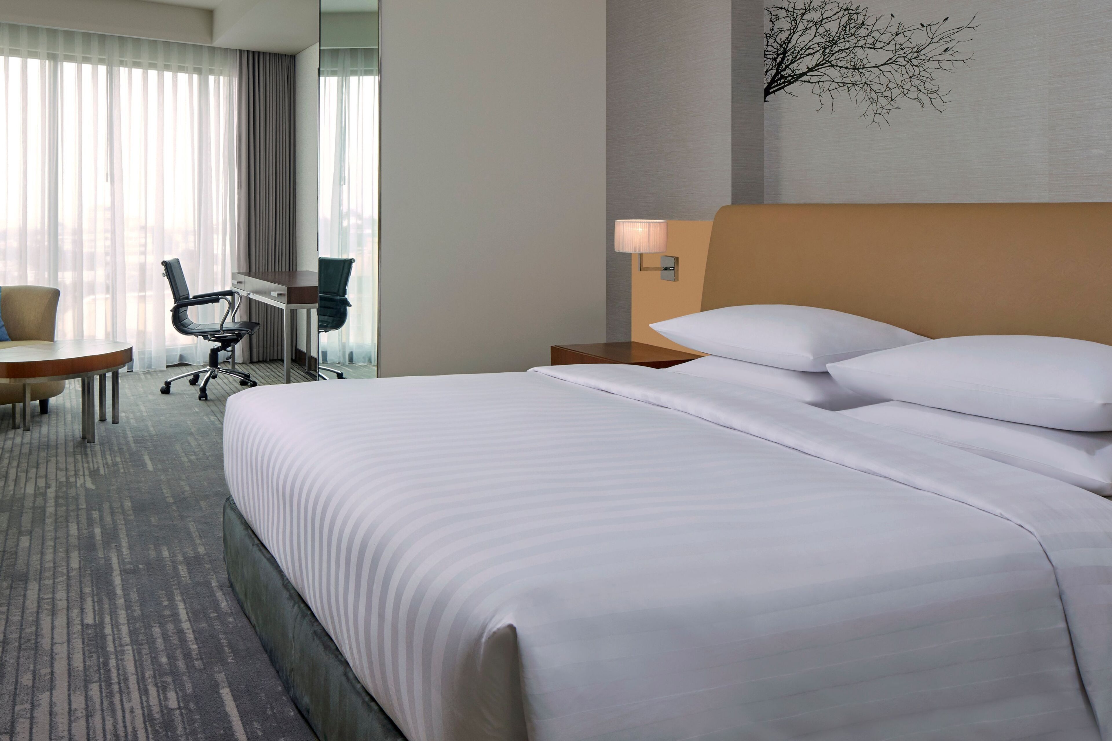 A hotel room with a large bed, a desk with a chair, and a view of the city through sheer curtains.