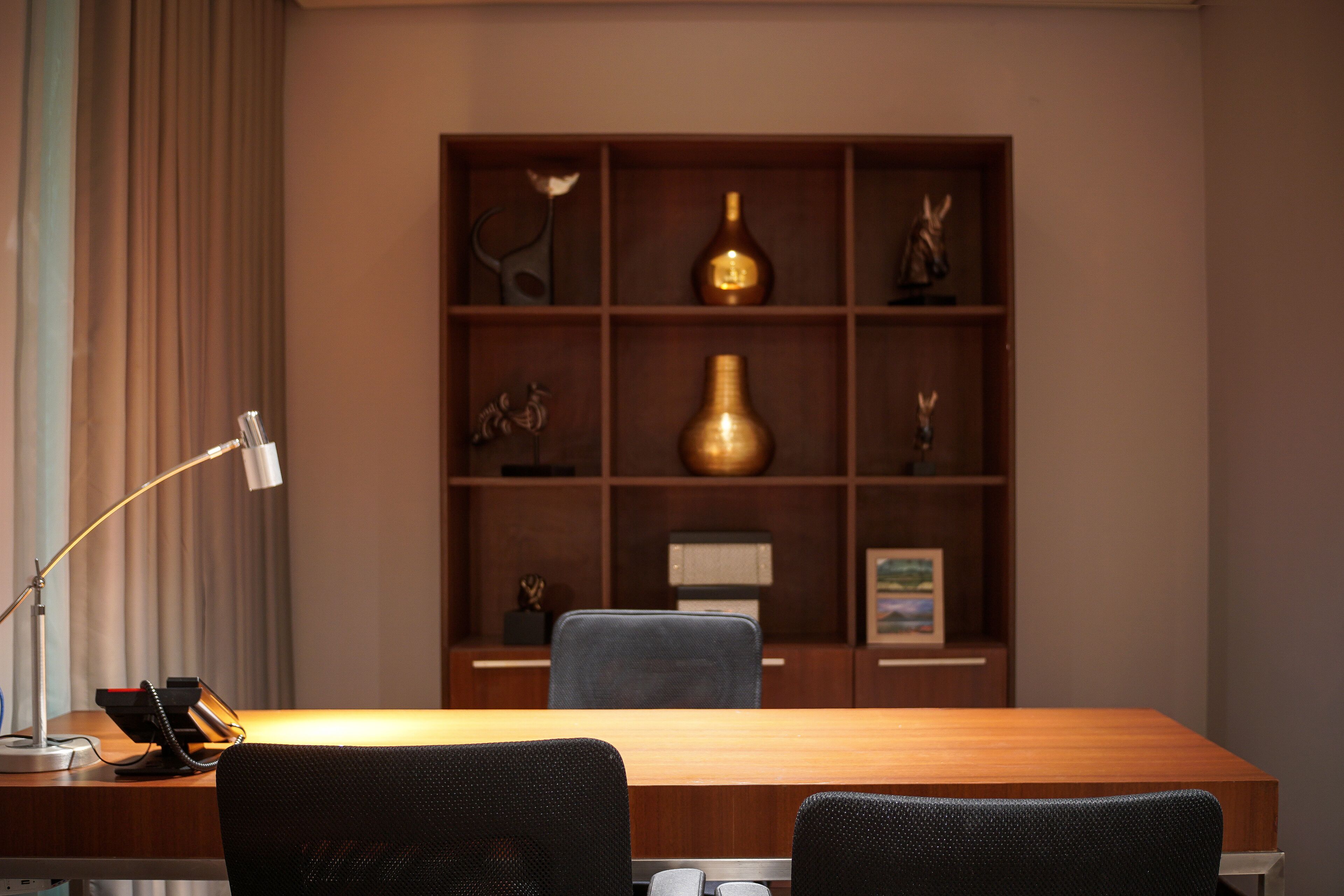 Una sala de conferencias con una mesa de madera, dos sillas, una lámpara de escritorio, un teléfono y una estantería decorativa con diversos objetos.