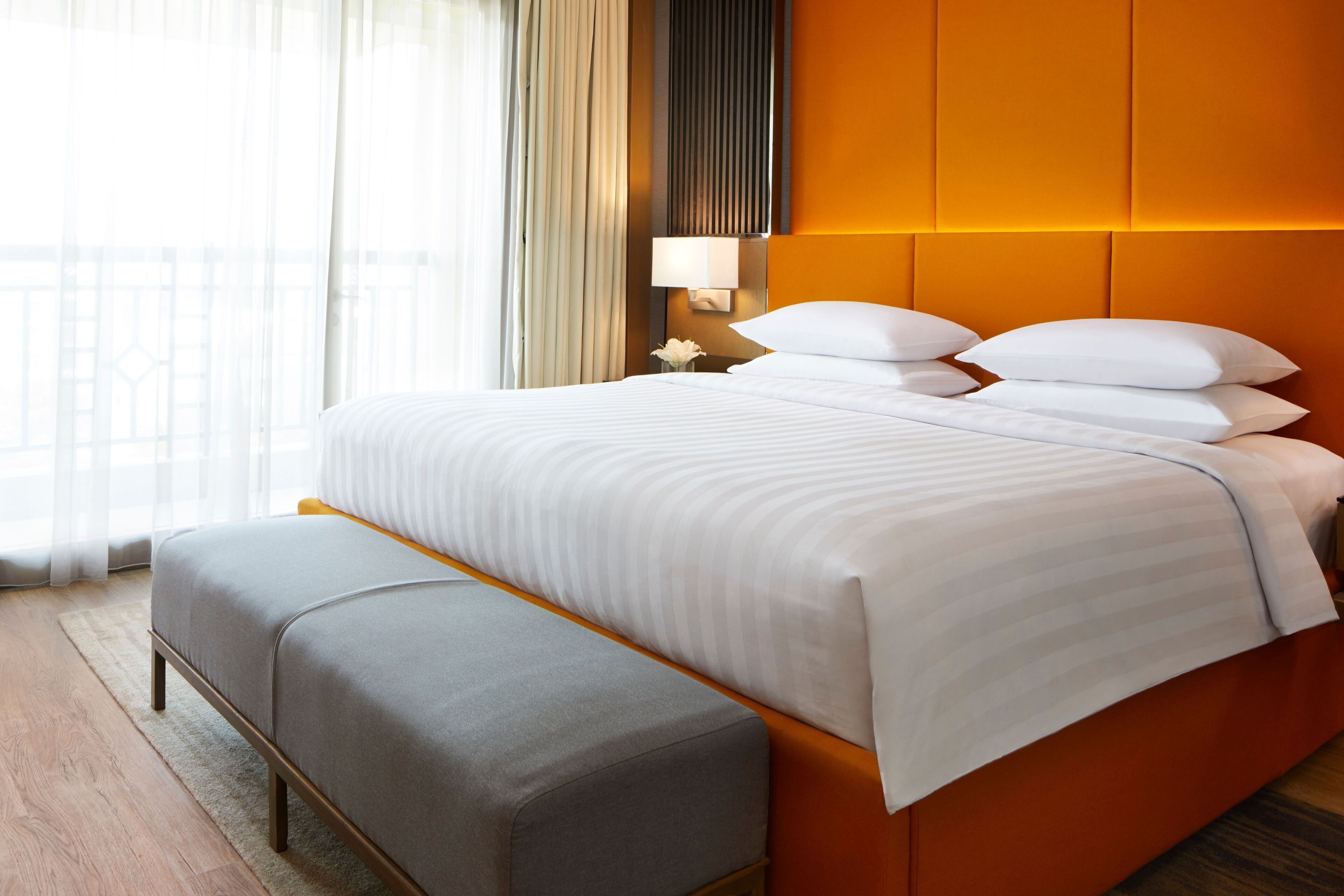 A hotel room with a large bed, a bedside table with a lamp, and a view of the outdoors through sheer curtains.