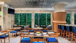 A modern restaurant with wooden tables and blue cushioned chairs, large windows with a view of greenery, and a unique wooden column structure. - Courtyard by Marriott Bandung Dago (Bandung)