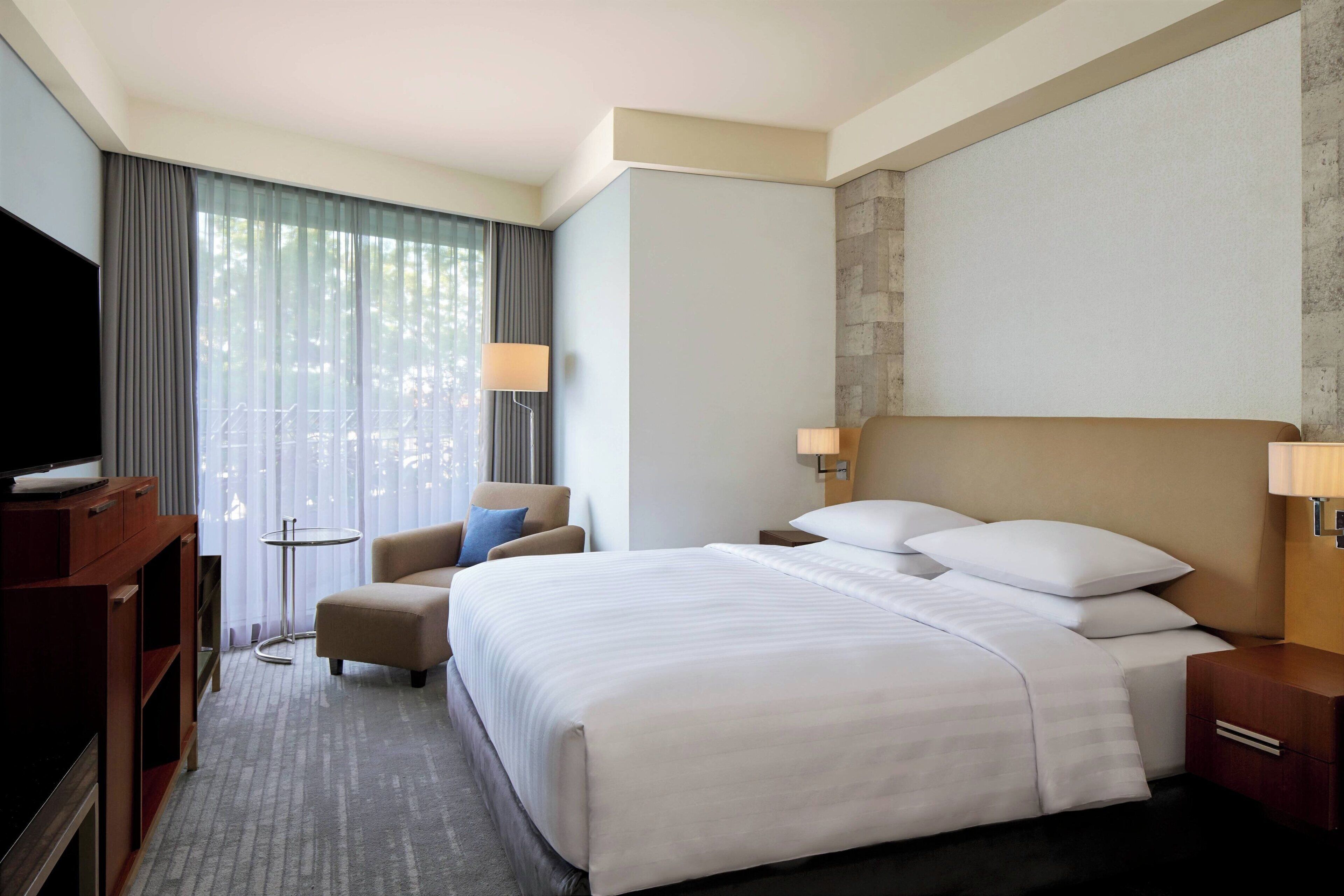 Habitación de hotel con una cama grande, televisor, una silla, una mesita y ventana con cortinas.