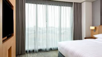 A hotel room with a large bed, a flat-screen TV, a window with sheer curtains, and a city view.