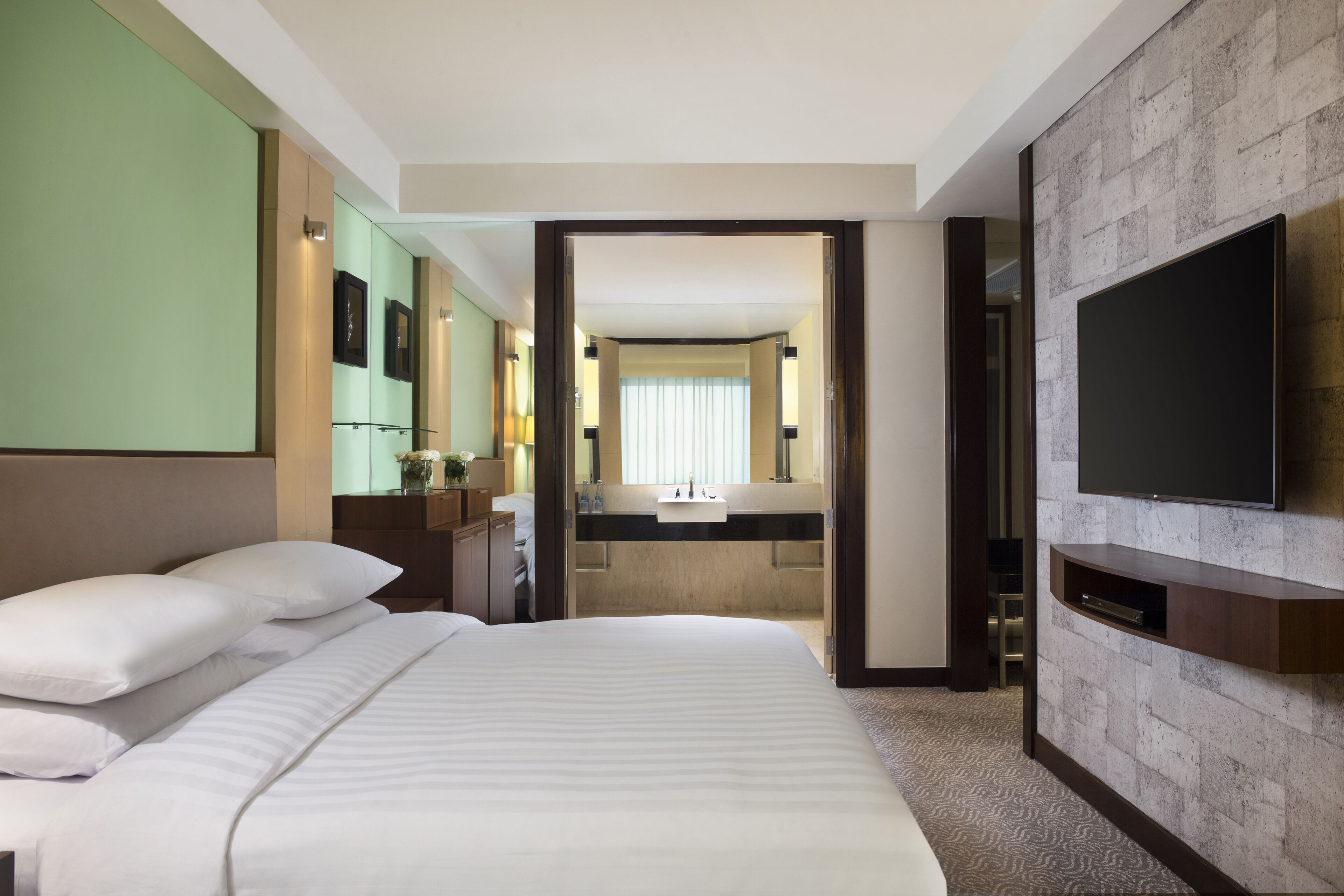 A hotel room with a large bed, a TV mounted on the wall, a bathroom with a mirror and sink, and a view into the hallway.