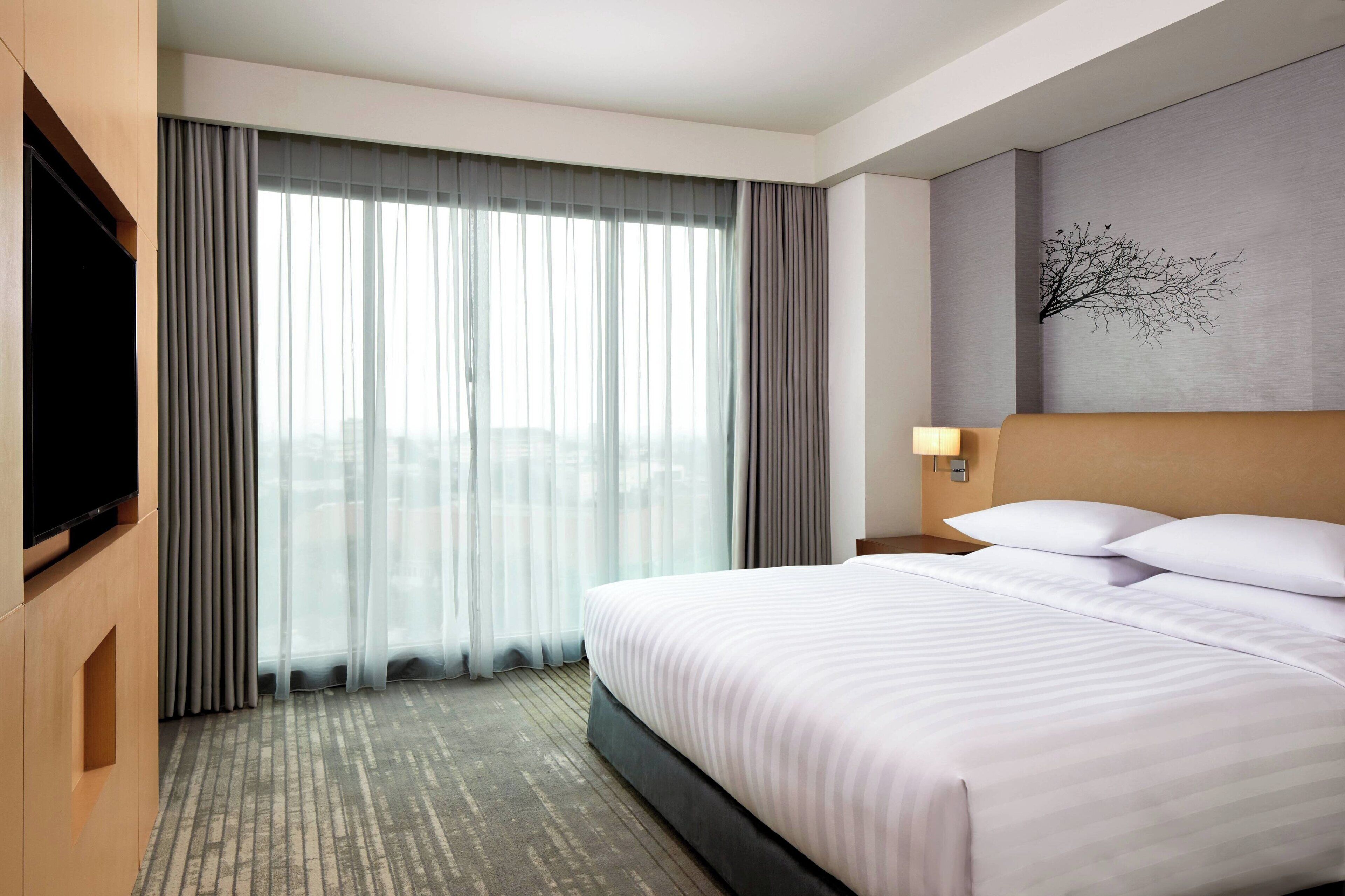 A hotel room with a large bed, a flat-screen TV, a wall-mounted lamp, and a window with sheer curtains.