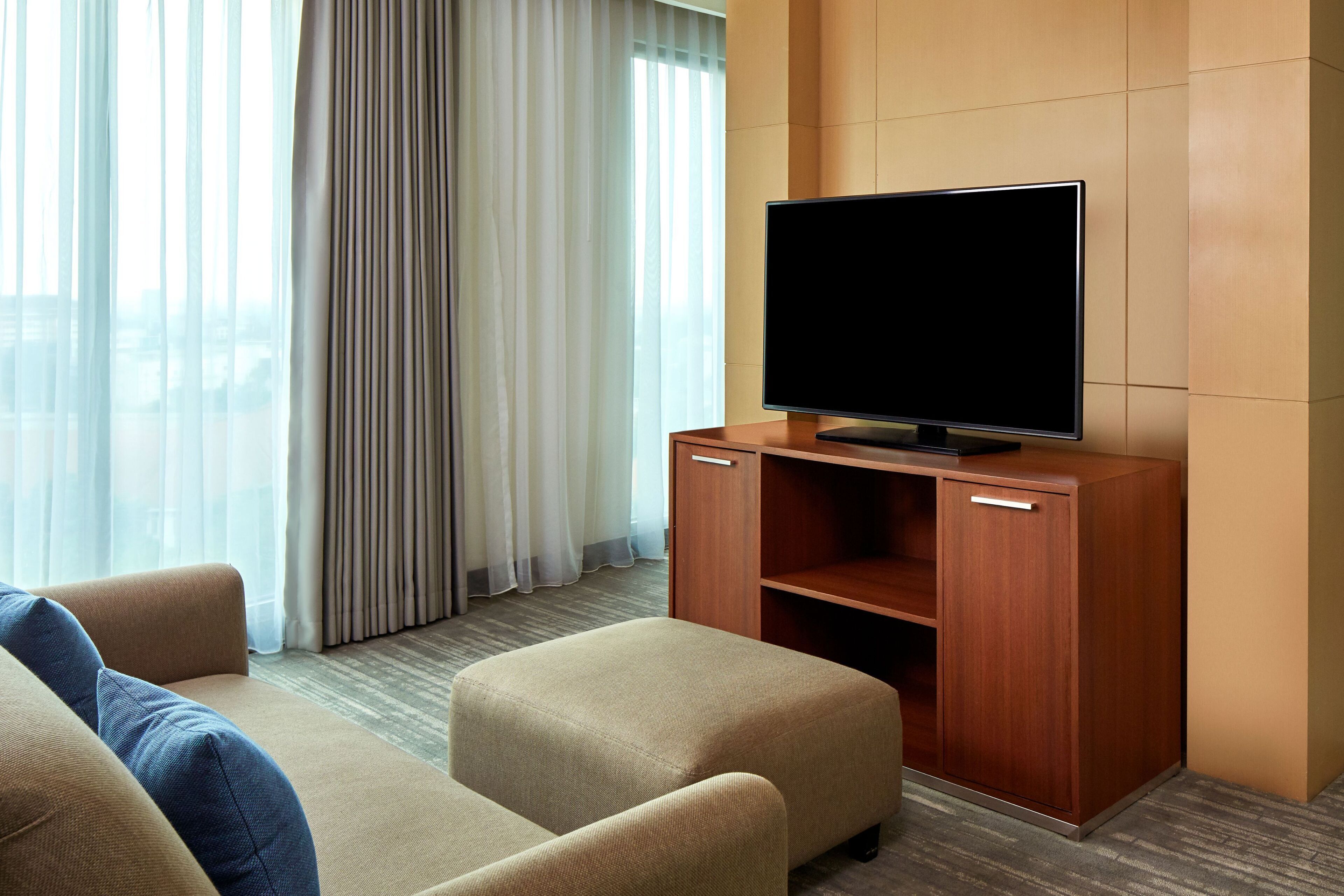 Ein modernes Hotelzimmer mit einem Flachbildfernseher auf einem Holzständer, einem beigen Sofa und einem großen Fenster mit transparenten Vorhängen.