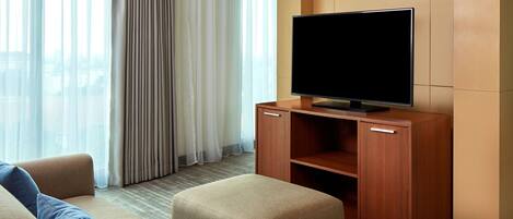 A modern hotel room with a flat-screen TV on a wooden stand, a beige sofa, and a large window with sheer curtains.