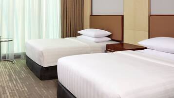 A hotel room with two beds, a small table, and a view of the outside through sheer curtains.