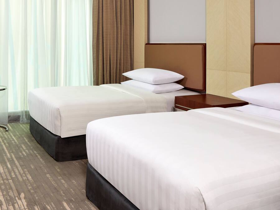 A hotel room with two beds, a small table, and a view of the outside through sheer curtains.