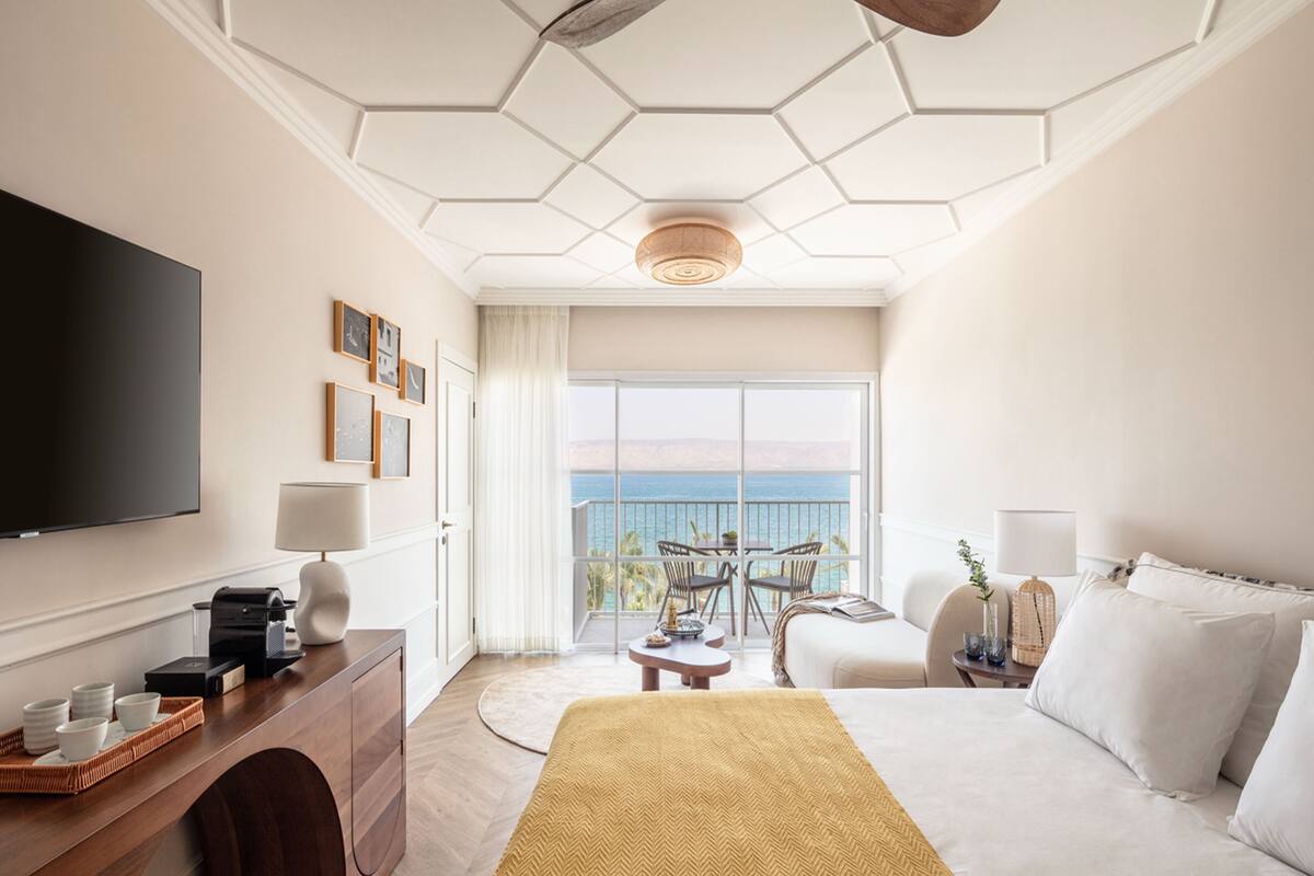 A hotel room with a large bed, a TV, a desk with a printer, a small table, and a balcony with a view of the sea.