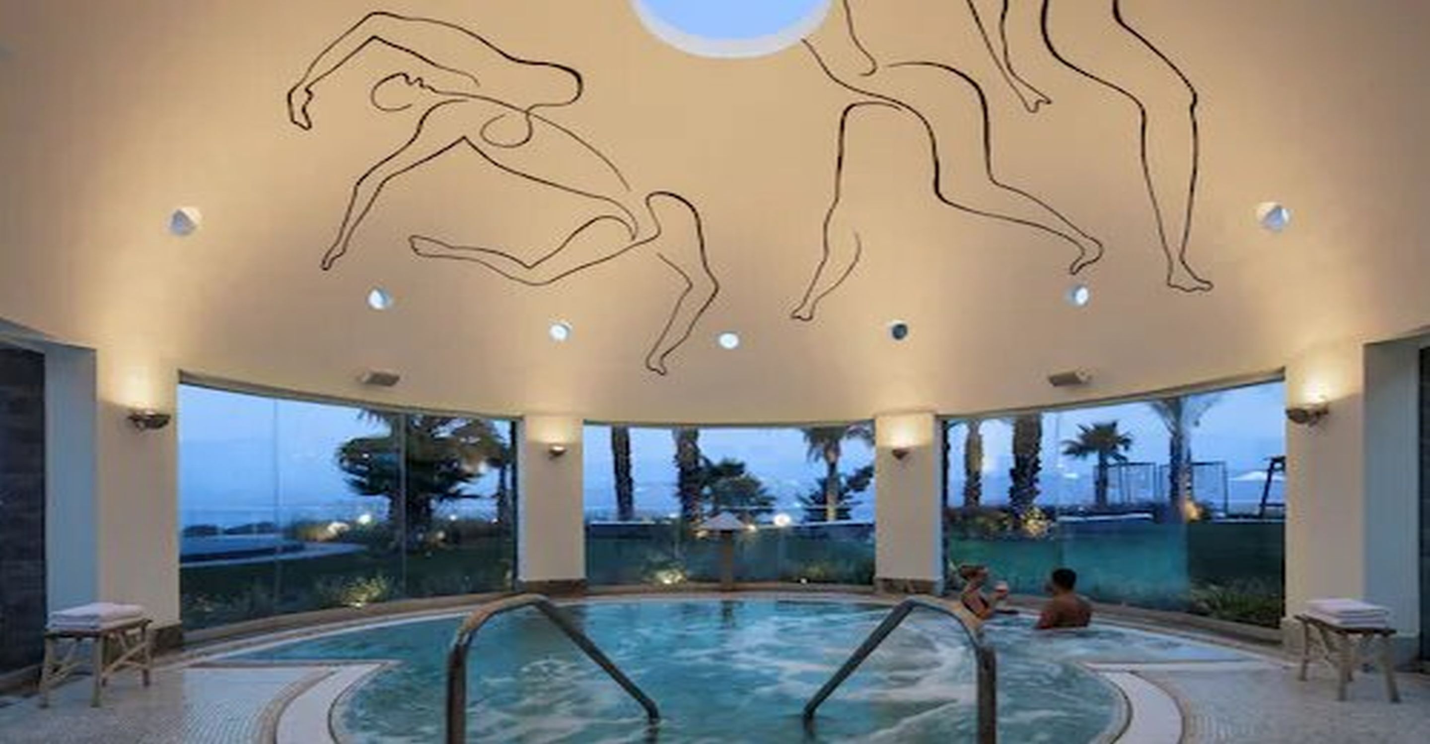 A circular indoor swimming pool with clear glass walls, allowing views of the outside. There are lounge chairs and a hot tub area.