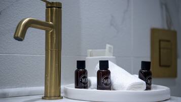 Un baño con grifo dorado, lavamanos blanco y tres botellas de productos para el cabello sobre una bandeja blanca.