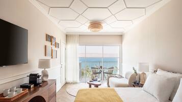A hotel room with a large bed, a TV, a desk with a printer, and a balcony with a view of the sea.